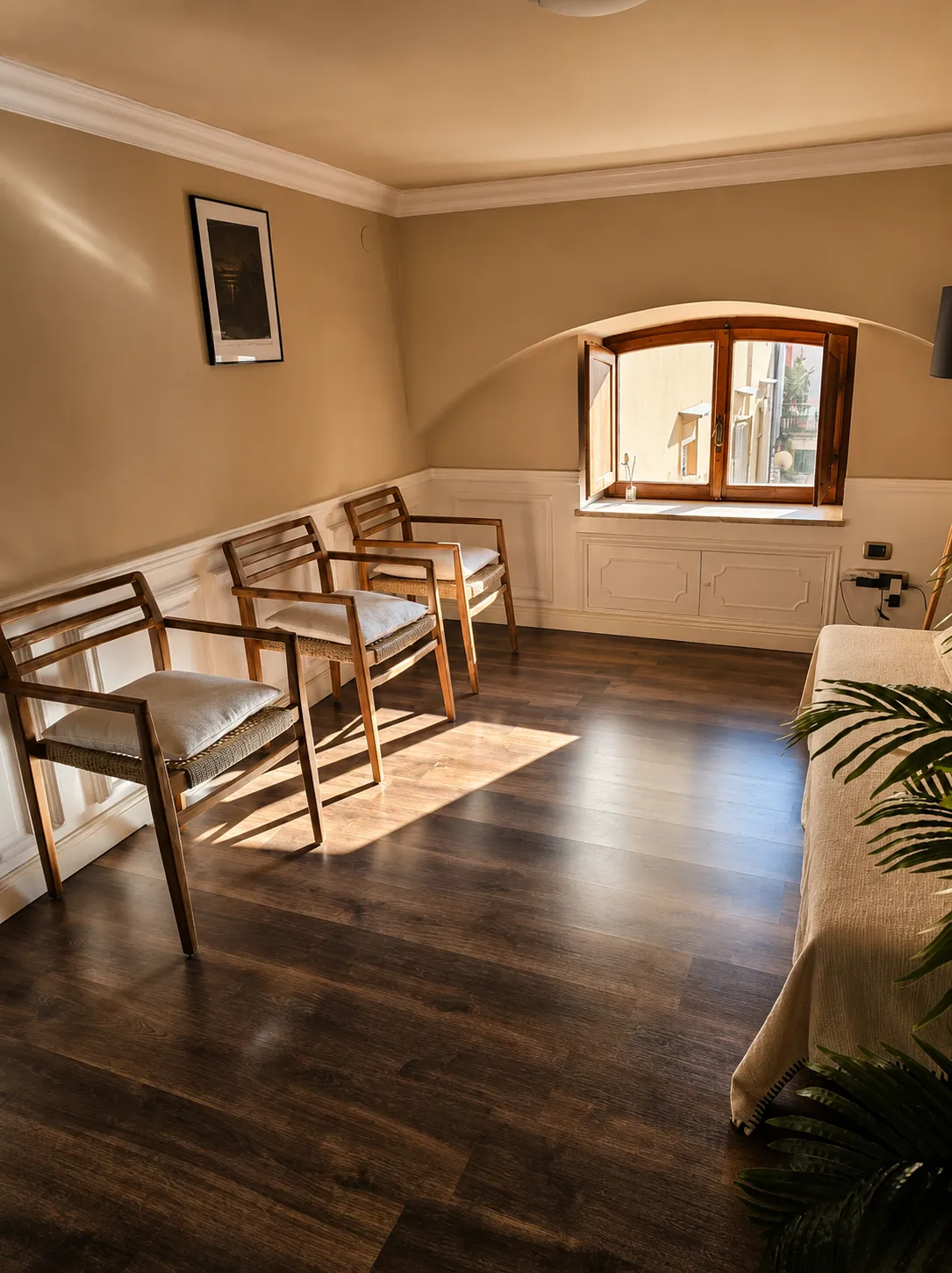 Sala d'attesa dello studio a Napoli, con sedie in legno, finestra ad arco, parquet scuro, divano chiaro e pianta.
